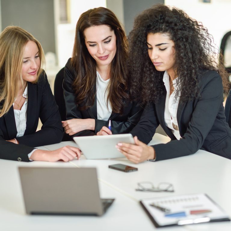 Three businesswomen working together in a modern office with white furniture. Teamwork concept. Caucasian, blonde, and muslim girls wearing suit. Multi-ethnic group of women